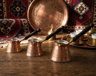 May include: Three copper coffee pots with wooden handles sit on a wooden table. The pots have intricate patterns and are accompanied by copper trays, cups, and a decorative plate. The background features patterned rugs and a copper plate.