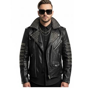Può includere: Giacca in pelle nera con borchie argentate su colletto, spalle e maniche. La giacca presenta un classico stile biker con revers a lancia, chiusura asimmetrica con zip e più tasche. È visibile una collana a catena argentata.