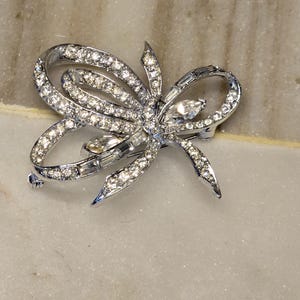 May include: A silver-toned metal brooch shaped like a looped ribbon or bow. The surface is encrusted with numerous small, clear, round-cut rhinestones and features larger, clear, marquise-cut stones set within the center and along the loops.
