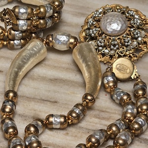 May include: An assortment of vintage jewelry, including a necklace with large, ivory-colored horn-shaped pendants and a bracelet with gold and silver beads. A large, ornate brooch with a central pearl and delicate floral detailing is also visible.