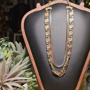 May include: A gold-toned necklace with two strands, one with a chain of interlocking oval links and the other a thinner snake chain. The necklace is displayed on a black jewelry stand with a wooden frame.