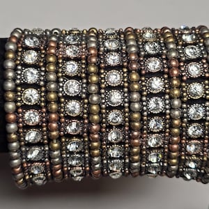 May include: A multi-strand bracelet featuring alternating rows of silver, bronze, and gold-toned beads. Clear, round rhinestones are set in square frames. The bracelet is a mix of metallic colors and sparkling accents.