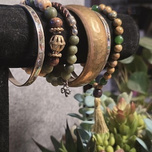 May include: A collection of bracelets displayed on a black stand. The bracelets include a silver bangle, beaded bracelets with green and brown beads, a hammered copper bangle, and a bracelet with a tassel charm. The background features greenery.