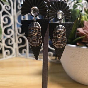 May include: A pair of black earrings with an Egyptian pharaoh design. The earrings feature a black fan-shaped top with a clear gem, a black triangle, and a silver-colored pharaoh pendant. The earrings are displayed on a brown stand.