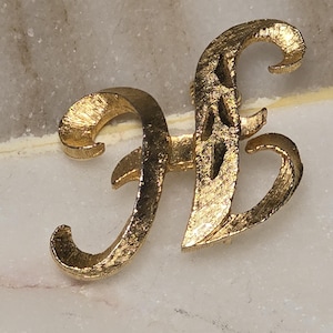 May include: A gold-toned metal brooch in the shape of the letter "H". The brooch has a textured surface and a decorative, cursive design. The pin is set against a light-colored, marble-like background.