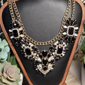 May include: A statement necklace with a gold chain and black and white gemstone accents. The necklace features multiple layers of chain and large, ornate pendants with a mix of black, white, and clear stones. The necklace is displayed on a black mannequin.