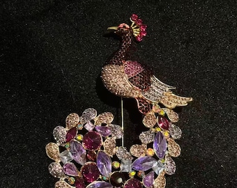 Puede incluir: Broche de pavo real enjoyada con un marco de metal dorado. El cuerpo del pavo real está adornado con cristales morados y transparentes, y las plumas de la cola son una mezcla de cristales morados, rosas y dorados. Sobre un fondo negro.