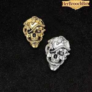 Puede incluir: Dos broches con forma de calavera, uno dorado y otro plateado, cada uno con una serpiente enrollada alrededor de la cabeza. Los broches tienen rasgos faciales detallados y están sobre un fondo oscuro. El texto "HerBroochBox" está en la esquina superior derecha.