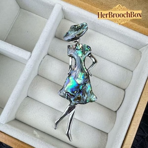 May include: Abalone shell brooch in the shape of a woman. The brooch has a silver-tone metal body and legs, with an iridescent abalone shell dress and hat. The brooch is displayed in a wooden box with a white interior. The text "HerBroochBox" is visible.