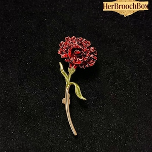May include: A red carnation flower brooch with green leaves and a gold stem. The brooch is set against a black background, with the text "HerBroochBox" in a gold frame.