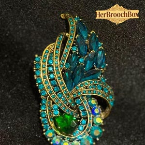 Könnte beinhalten: Eine verzierte Brosche mit goldfarbenem Rahmen, verziert mit türkis- und grünen Kristallen. Das Design zeigt wirbelnde Bänder und eine zentrale Ansammlung türkisfarbener Steine. Der Text "HerBroochBox" ist oben rechts sichtbar.