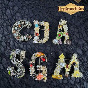 Puede incluir: Un surtido de broches de letras enjoyadas que deletrean "CDA SGM". Cada letra está incrustada con gemas de colores, perlas y cristales, engastados en metal dorado. Los broches se exhiben sobre una superficie negra texturizada.