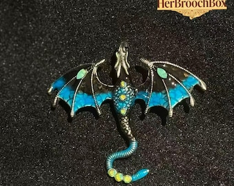 Puede incluir: Broche de dragón con metal plateado, esmalte negro y turquesa. El dragón tiene alas turquesas con detalles en negro y detalles de gemas amarillas y verdes. El broche tiene el texto "HerBroochBox".