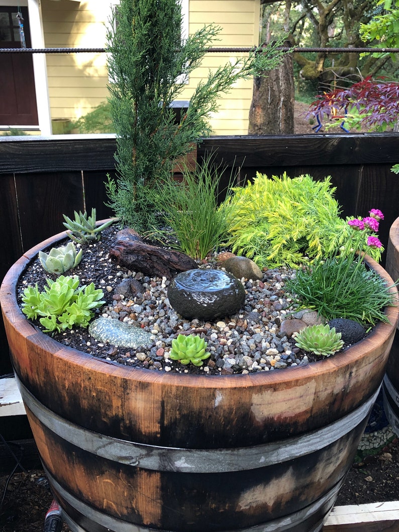 May include: A wooden barrel planter filled with various succulents, small plants, and decorative stones. A small, dark stone fountain is in the center. The barrel has metal bands and is set outdoors.
