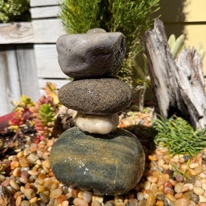 May include: A balanced stone sculpture composed of four smooth, grey and tan rocks of varying sizes. The sculpture is wet and rests on a bed of small, colourful pebbles. Green plants and wood are in the background.