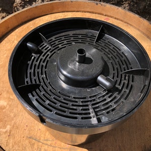 May include: A black plastic wine barrel insert with a circular design and a central raised section. The insert has a series of curved slots and a small spout. It sits on a wooden barrel.