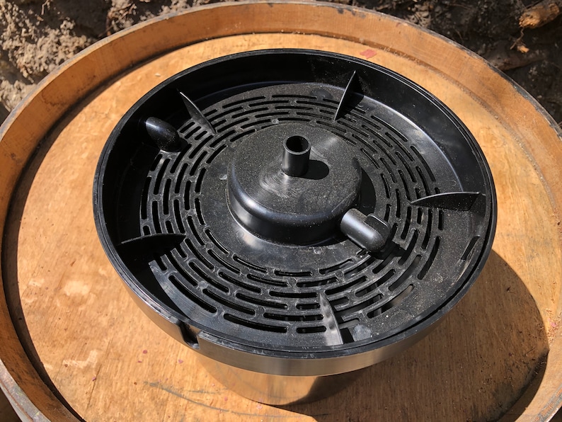 May include: A black plastic wine barrel insert with a circular design and a central raised section. The insert has a series of curved slots and a small spout. It sits on a wooden barrel.