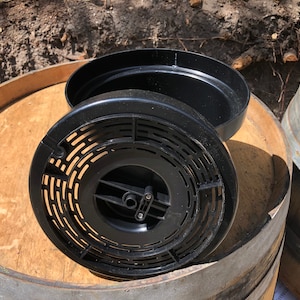 May include: Black plastic component with a circular, vented design, possibly a part of a larger system. The component is open, revealing its interior structure. It rests on a wooden surface, likely a barrel top.