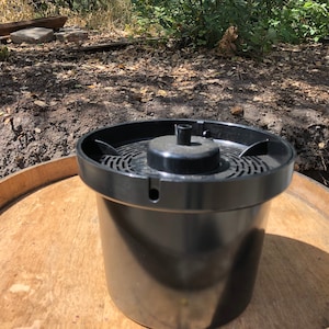 May include: A black plastic film developing tank with a perforated top and a central spindle. The tank is cylindrical and sits on a wooden surface, likely a barrel top. The background shows a natural outdoor setting.