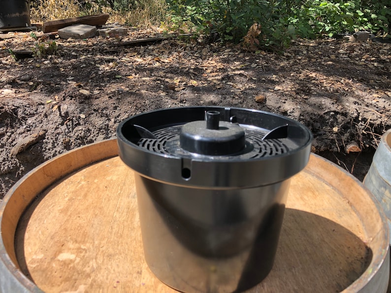 May include: A black plastic film developing tank with a perforated top and a central spindle. The tank is cylindrical and sits on a wooden surface, likely a barrel top. The background shows a natural outdoor setting.