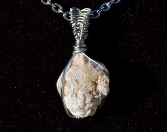 May include: A handcrafted pendant necklace featuring a light beige, textured stone wrapped in silver-colored wire. The pendant hangs from a delicate silver chain, creating a unique and natural accessory. The necklace is displayed against a dark background.