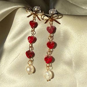 May include: Dangling earrings with a gold bow design at the top. Each earring features three red heart-shaped beads connected by a gold chain, and a white pearl at the bottom. The earrings are set against a cream-colored background.