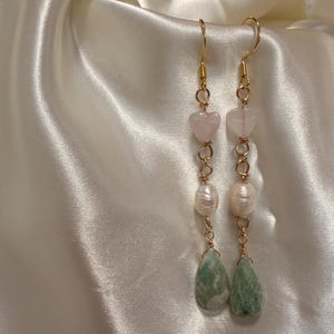May include: Gold-toned dangle earrings featuring heart-shaped pink stones, white pearls, and teardrop-shaped green stones. The earrings are suspended from gold-colored ear wires and connected with delicate gold chain links. The earrings are displayed on a white satin background.