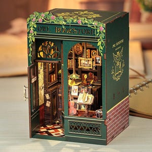May include: A miniature Owl Bookstore diorama with a detailed interior, including books, a guitar, and framed pictures. The exterior is green with the words "OWL BOOKSTORE" and a brick facade. The diorama is illuminated with warm lighting.
