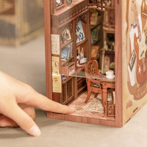May include: A miniature wooden bookshelf diorama with a detailed interior, including a desk, chair, and books. The diorama features a warm, inviting color palette with intricate details, creating a cozy, vintage aesthetic. A hand is pointing at the diorama.