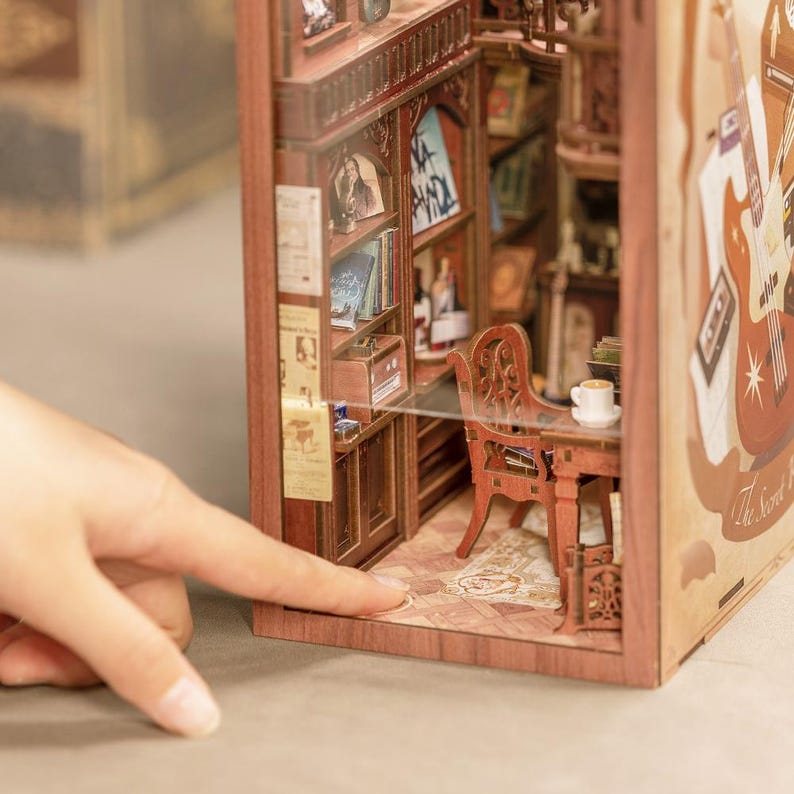 May include: A miniature wooden bookshelf diorama with a detailed interior, including a desk, chair, and books. The diorama features a warm, inviting color palette with intricate details, creating a cozy, vintage aesthetic. A hand is pointing at the diorama.
