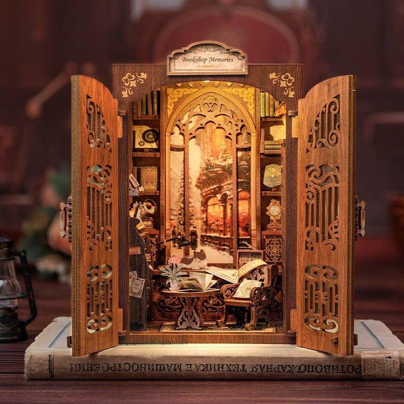 May include: A miniature wooden bookshelf diorama with openwork doors, featuring a detailed interior scene of a cozy reading nook. The diorama includes a table, chair, books, and a warm, inviting glow from the interior lighting. The sign on top reads "Bookshop Memories".