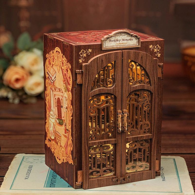 May include: A wooden decorative box with a hinged door design. The box is brown with intricate cut-out patterns and a small sign that reads "Bookshop Memories". The side of the box features a detailed illustration in shades of orange and gold.
