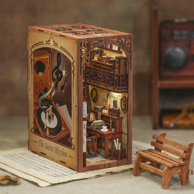 May include: A wooden book nook with a detailed interior scene. The front features a record player and musical notes, with the text "The Secret Rhythm." Inside, a miniature room with a desk, chair, and balcony is visible. A small wooden bench sits in front.