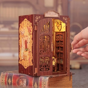 May include: A miniature wooden bookshelf model with an open door revealing a detailed interior. The model is brown with intricate carvings and a decorative facade. The text "Bookshop Memories" is above the door. The model is placed on top of a large book.