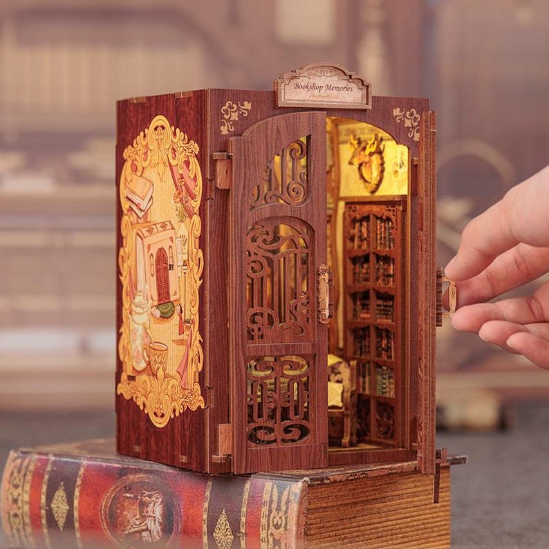 May include: A miniature wooden bookshelf model with an open door revealing a detailed interior. The model is brown with intricate carvings and a decorative facade. The text "Bookshop Memories" is above the door. The model is placed on top of a large book.