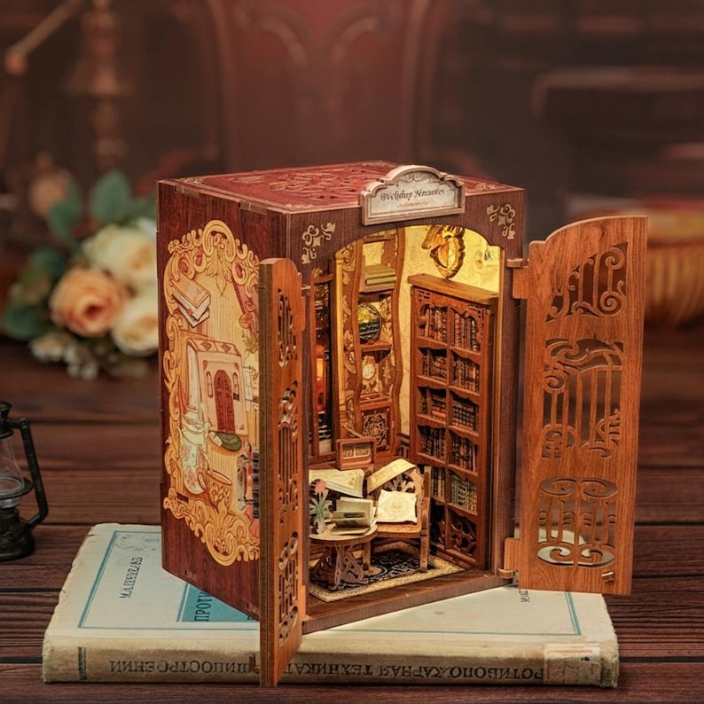 May include: A wooden miniature bookshelf with open doors, revealing a detailed interior scene of a library. The exterior is decorated with ornate carvings and a small sign. The miniature is set on top of a book.