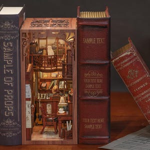 May include: A miniature wooden bookshelf diorama with a detailed interior, featuring a cat on a balcony overlooking a Parisian scene. Flanking the diorama are two faux books with the text "SAMPLE OF PROPS" and "SAMPLE TEXT". The diorama includes a desk, chair, and other miniature furniture.