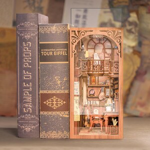 May include: A wooden bookshelf insert with a miniature room scene, featuring a cat on a balcony, books, and furniture. Flanking the insert are two decorative book covers with text: "SAMPLE OF PROPS" and "TOUR EIFFEL".
