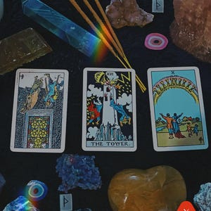 May include: A collection of crystals, tarot cards, and incense sticks arranged on a black surface. The tarot cards include "The Tower" card. Various crystals are visible, including a blue quartz point and a heart-shaped citrine.