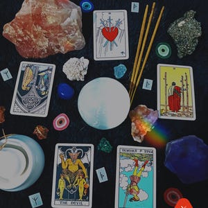 May include: An assortment of crystals, tarot cards, incense, and decorative items arranged on a dark surface. The tarot cards feature various illustrations, including a heart pierced by swords and a devil figure. A rainbow prism effect adds color.