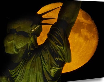 May include: A canvas print of the Statue of Liberty silhouetted against a large, orange-yellow moon. The statue is a dark green color, with the moon providing a warm glow in the background. The artwork is set against a black background.