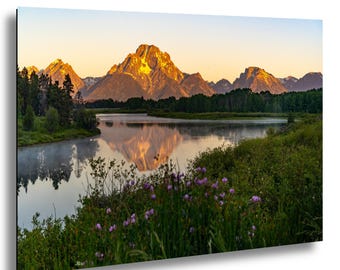 Puede incluir: Impresión de un paisaje montañoso al amanecer. Las montañas están iluminadas con luz dorada, reflejada en el agua tranquila. La exuberante vegetación verde y las flores silvestres moradas enmarcan el primer plano.