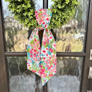 May include: A green wreath hangs on a dark brown door. A floral scarf with pink and red flowers on a light blue background is tied to the wreath. The door has glass panels.