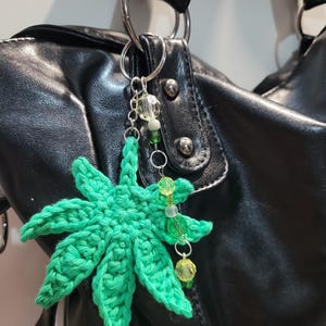 May include: A green crocheted cannabis leaf keyring with a silver chain and decorative beads. The keyring is attached to a black leather handbag. The leaf is a vibrant green colour and has detailed stitching.