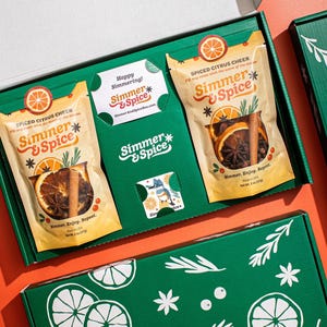 May include: A green gift box containing two beige pouches labeled Simmer & Spice Spiced Citrus Cheer, each holding dried orange slices, cinnamon sticks, and star anise. The box includes a small card reading Happy Simmering and Simmer & Spice.