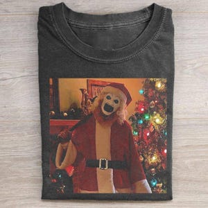 May include: Dark gray t-shirt featuring a creepy Santa Claus figure with a white mask and red Santa suit. The image shows the figure in front of a Christmas tree with colorful lights. The shirt is made of a soft material.