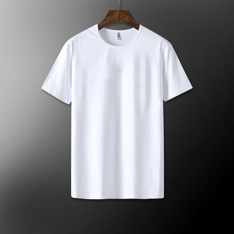 May include: A plain white crew neck t-shirt hanging on a wooden hanger. The short-sleeved shirt is made of a smooth, opaque fabric. The background is a gradient of gray and black.