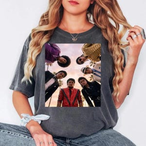 May include: Dark gray t-shirt featuring a group of people in a circle, with a sunset background. The people are wearing various outfits, including a red jacket, black leather jacket, and suits. Palm trees are visible in the background.
