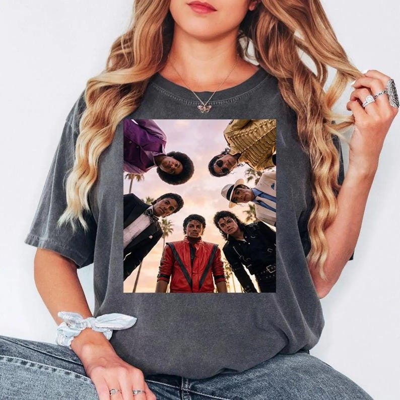May include: Dark gray t-shirt featuring a group of people in a circle, with a sunset background. The people are wearing various outfits, including a red jacket, black leather jacket, and suits. Palm trees are visible in the background.