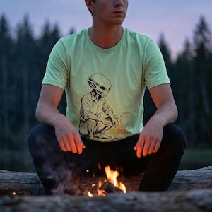 May include: Light green t-shirt featuring a detailed illustration of an alien sitting by a campfire. The alien is depicted in a seated position, wearing a button-down shirt. The shirt is a soft, comfortable material.